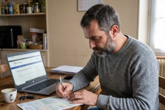 Homme remplissant un formulaire de demande de carte grise à la maison avec un stylo et des documents officiels sur une table en bois
